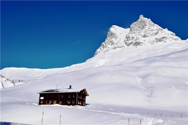 A cozy mountain hut in the snow in front of an impressive mountain backdrop. The clear blue sky creates a beautiful winter atmosphere.