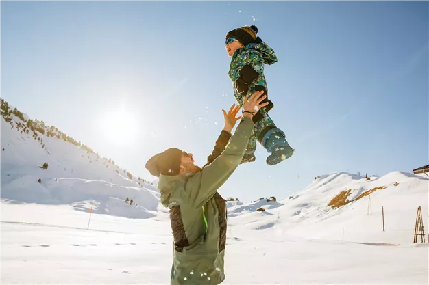 A father lifts his child into the air, surrounded by snow-covered mountains. The sun shines brightly in the blue sky.
