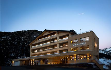 Ein modernes Hotelgebäude in den Bergen, umgeben von schneebedeckten Landschaften. Der Himmel zeigt sanfte Dämmerungsfarben.