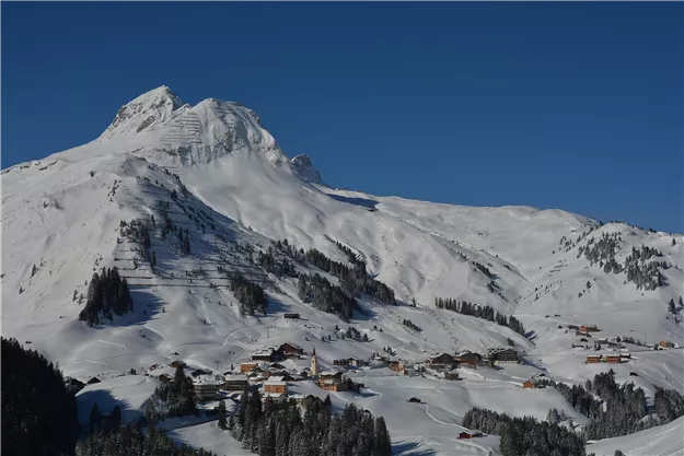 Eine schneebedeckte Berglandschaft unter einem klaren blauen Himmel. Im Vordergrund ist ein kleines Dorf mit traditionellen Häusern zu sehen.