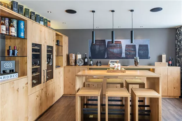 A modern room with a wooden bar and chairs. In the background, there are shelves with bottles and a stylish wall design.