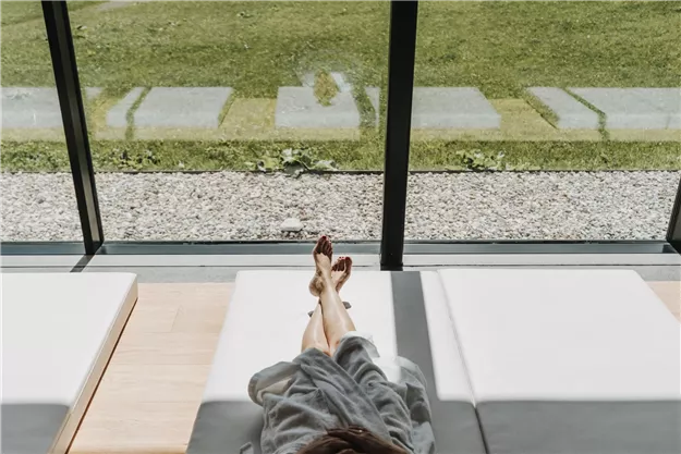 A person relaxes on a lounge chair with a view of a well-kept garden. The large window front lets in a lot of light.