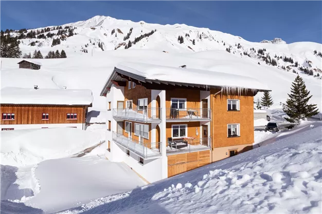 A charming wooden house in the snow with balconies and surrounding mountains. The entire landscape is wrapped in a peaceful blanket of snow.
