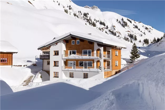 A beautiful, snow-covered Alpine house with wood and glass details. In the background, snow-covered mountains can be seen.
