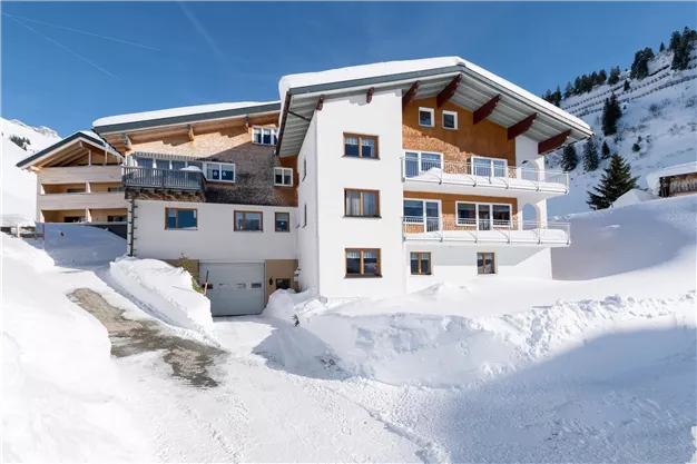 A modern building in the snow with balconies and a clear blue sky. The surroundings are covered with fresh snow, showcasing an alpine landscape.