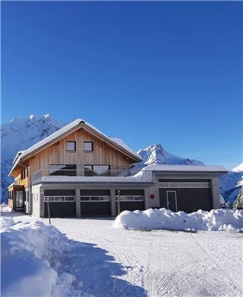 Ein modernes Haus in einer Schneelandschaft mit Bergen im Hintergrund. Der Himmel ist klar und blau, und der Weg ist schneebedeckt.