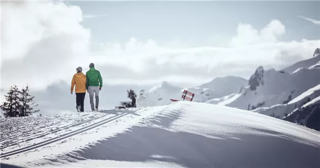 Zwei Menschen gehen auf einem schneebedeckten Weg in den Bergen. Die Landschaft ist malerisch mit schneebedeckten Gipfeln und klarem Himmel.