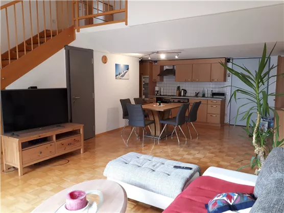 A cozy living room with a modern kitchen and a dining table. The wooden staircase leads to the upper area of the room.