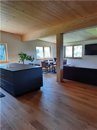 A modern apartment with an open living area and wooden floor. There is a table with chairs by the window and a plant is visible.