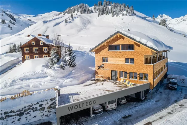 A modern wooden building in the snow with a sign that says "WOLFEGG." In the background, snow-covered mountains and other alpine huts are visible.