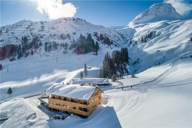 A picturesque mountain landscape with snow-covered hills and a cozy wooden house. The clear blue sky creates a dreamy winter atmosphere.