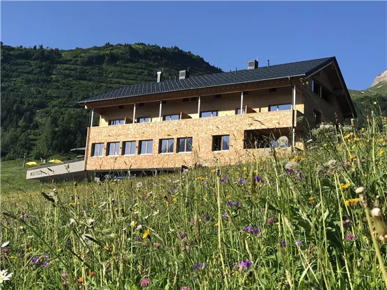 A modern building in the mountains with many windows.
Surrounded by a colorful meadow full of flowers and green hills.