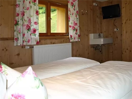 Ein gemütliches Schlafzimmer mit zwei Betten und Holzwänden. Die Fenster sind mit blumigen Vorhängen dekoriert und ein kleiner Fernseher ist an der Wand montiert.