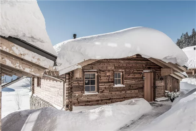 Eine gemütliche Blockhütte in einer schneebedeckten Umgebung. Der Schnee liegt hoch auf dem Dach und strahlt eine winterliche Atmosphäre aus.